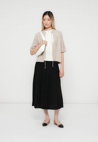 Hellbrauner, taillierter Cardigan mit Kordelzug, weißem, figurbetontem Top, schwarzem, plissiertem Midirock und braunen Ballerinas mit schwarzen Spitzen, die eine weiße Tasche halten.