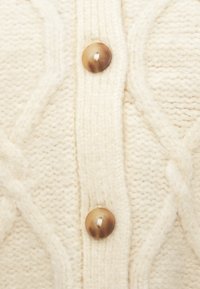 Πλεκτό ύφασμα σε κρεμ απόχρωση με σχέδια κοτσίδας και δύο στρογγυλά καφέ κουμπιά ραμμένα κάθετα στο κέντρο.