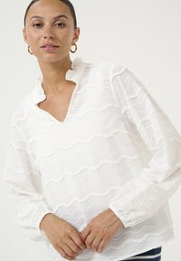 Blusa bianca con pattern a onde stratificate, tessuto testurizzato, colletto con frange e maniche elastiche. Presenta piccoli pompon lungo le cuciture.