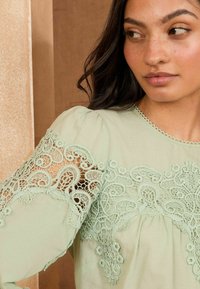 Blusa verde chiaro con dettagli in pizzo sulla manica, scollatura rotonda con bordo scallop e morbida trama del tessuto.
