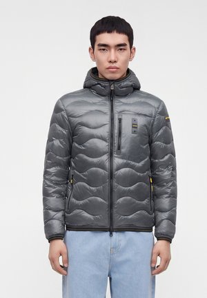 Jeune homme portant une veste matelassée grise à capuche avec poches zippées à l'avant et sur la poitrine, associée à un jean bleu clair, debout devant un fond uni.