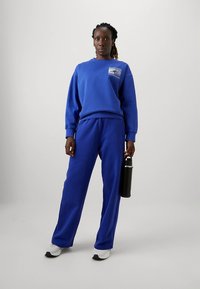 Blue cotton sweatshirt and matching pants with a relaxed fit, featuring a logo patch on the chest. A black water bottle is held in one hand.