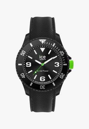 Musta aurinkovoimalla toimiva kello, jossa on teksturoitu kumiranneke, pyöreä musta kellotaulu, valkoiset tuntimerkit, kolme viisaria ja vihreä yksityiskohta kruunussa.