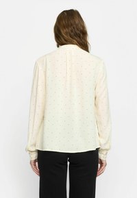 Blusa a maniche lunghe di colore crema con piccoli pois neri, tessuto leggero, polsini con bottoni e orlo arrotondato. Senza motivi o dettagli distintivi.