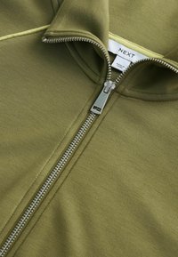Vêtement vert olive avec fermeture éclair en métal argenté partiellement ouverte, montrant une étiquette blanche avec la marque "NEXT" et le texte "Made in China".