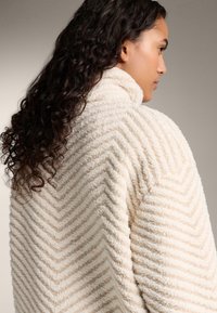 Cremfarbener, strukturierter Mantel mit Zickzackmuster, der einen hohen Kragen und ein weiches, plüschiges Material aufweist; lockige Haare teilweise sichtbar.