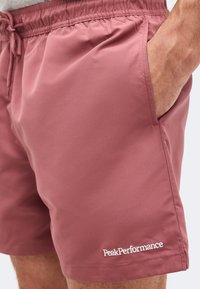 Κοντινό πλάνο ατόμου που φοράει ροζ σορτς με ελαστική μέση, κορδόνι, πλαϊνή τσέπη και λευκό λογότυπο "Peak Performance" κοντά στο τελείωμα.