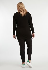Tunique noire texturée à manches longues avec un ourlet arrondi, associée à des leggings noirs et des baskets blanches avec des accents verts.