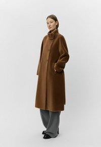 Brauner Wollmischmantel mit hohem Kragen, vorderen Knöpfen und Seitentaschen; kombiniert mit weiten grauen Hosen und schwarzen Schuhen.