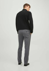Schwarzer Pullover mit hohem Kragen, kombiniert mit grauen Hosen. Die Hosen haben seitliche Taschen und eine Gesäßtasche mit einem Knopfdetail.