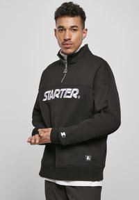 Svart halv-zip genser laget av mykt stoff, med stor hvit "STARTER" logo over brystet og logo aksenter på ermene.