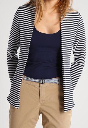 Kvinde iført en navy tanktop, navy og hvide stribet blazer, beige bukser og mønstret bælte mod en ensfarvet lys baggrund.