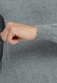 Manche de pull en tricot gris avec un poignet côtelé, montrant une main en position de poing. La texture semble douce et lisse, sans motifs visibles.