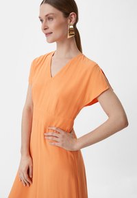 Pomarańczowa sukienka z dekoltem w serek, krótkimi rękawami, gładką teksturą, dopasowanym gorsetem i zwiewną spódnicą, podkreślona złotą geometryczną bransoletką.
