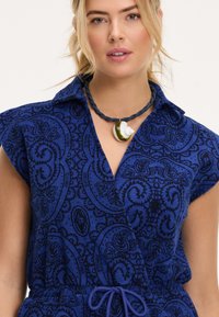 Mėlyna paisley rašto suknelė su trumpomis rankovėmis, apykakle ir juosmens diržu. Kaklo papuošalas su apvaliu pakabuku, kuriame yra žalių ir baltų akcentų.