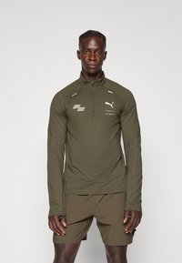 Olivengroen sportieve pullover met ritskraag, lange mouwen, reflecterende accenten en logo op de borst; gecombineerd met bijpassende shorts.