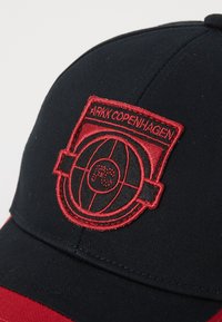 Black cap with red accents, featuring an embroidered patch displaying a circular emblem and the text "ARKK COPENHAGEN." Smooth fabric texture.