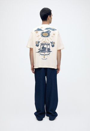 Hombre de espaldas lleva una camisa corta de color crema con sol, nubes, pájaros y elementos arquitectónicos, combinada con pantalones anchos azul oscuro.