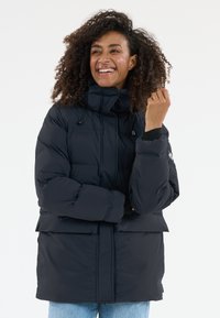Lächelnde Frau in einer schwarzen Steppjacke und blauen Jeans, mit lockigem Haar vor einem schlichten weißen Hintergrund.