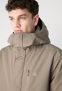 Beige hettejakke med høy krage, som har en glidelås-lomme og en logo på brystet. Laget av et glatt og lett materiale.