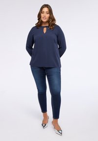 Top de manga larga azul marino con escote de ojal y tela texturizada, combinado con vaqueros ajustados de lavado oscuro y tacones plateados de punta.