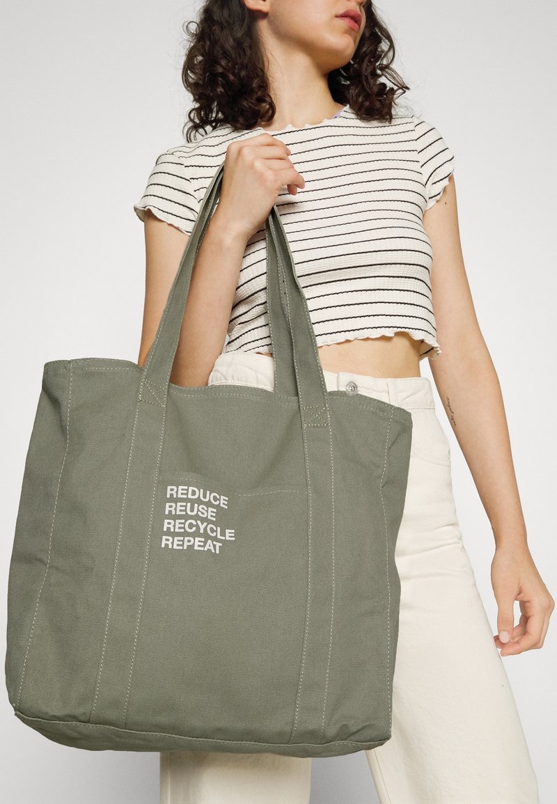 Femme portant un crop top rayé tenant un grand sac cabas vert avec le texte «REDUCE REUSE RECYCLE REPEAT» imprimé en blanc.