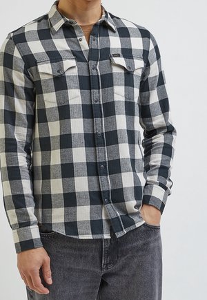 Hombre con camisa de cuadros en blanco y negro con bolsillos en el pecho y jeans gris oscuro, con una mano en el bolsillo.