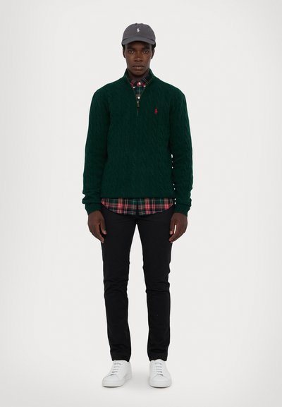 Dark green cable-knit sweater with zip collar, red logo. Layered over checkered plaid shirt, black pants, and white sneakers. Gray cap.