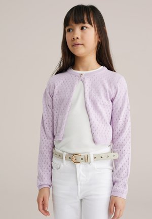 Jeune fille portant un cardigan tricoté violet clair sur un haut blanc, un pantalon blanc et une ceinture beige avec des découpes en forme de cœur.