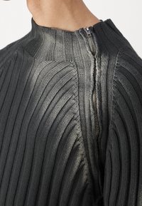 Grå ribbad stickad tröja med hög krage och en sned dragkedja. Texturen är fint detaljerad med vertikala linjer.