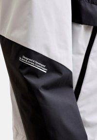 Čierno-biely vetrovka s elegantným dizajnom, hladkou textúrou a logovou značkou na rukáve. Zapínanie na zips vpredu.