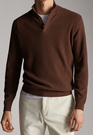 Mand iført en brun kvart-zip sweater over en hvid undertrøje kombineret med off-white teksturerede bukser mod en ensfarvet baggrund.