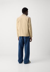 Beige lichtgewicht jas met een strak design, voorzien van een kraag en elastische manchetten. Gecombineerd met los zittende blauwe denim jeans.