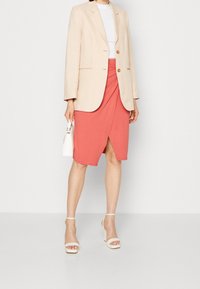 Beige puku na mabuttoni mawili, koti la ndani la buluu lililo na muundo mzuri, sketi ya mwavuli ya coral, na viatu vya beige vyenye mikanda. Anashikilia mkoba mdogo mweupe.