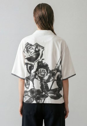 Osoba s mokrými vlasy, která má na sobě bílou krátký rukáv košili s velkým černým vertikálním pruhovaným růžovým vzorem na zádech.