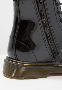 Botines de charol negro con punta redonda, cremallera lateral, costura en contraste amarilla en la suela y tacón texturizado estriado.