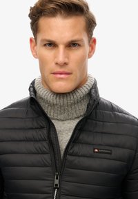 Chaqueta de plumas negra con patrón acolchado, cierre de cremallera y cuello alto, llevada sobre un suéter de cuello alto de canalé gris. Logo de la marca en el pecho.