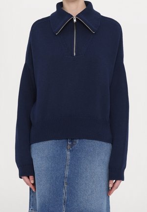 Femme portant un pull en maille bleu marine avec un col zippé à l'avant et une jupe en jean bleue.