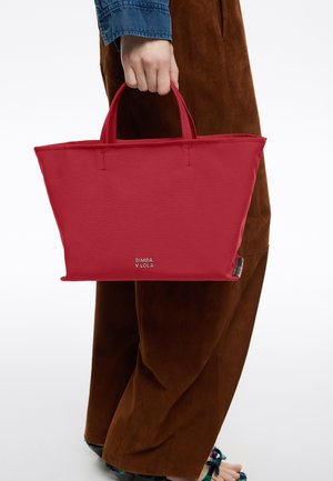 Czerwona torba na ramię wykonana z materiału o gładkiej fakturze, z krótkimi uchwytami i logo na dole. Minimalistyczny design, prostokątny kształt.