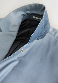 Col de chemise bleu clair avec un bouton caché et une doublure intérieure noire, partiellement ouvert sur un fond neutre.