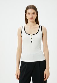 Hvid, ribbet tanktop med sorte detaljer, der har en rund halsudskæring og tre dekorative knapper foran. Glat stofstruktur.