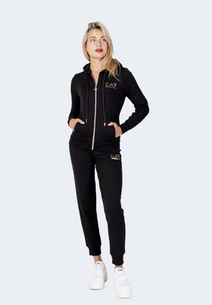 Haut à glissière noir et pantalon assorti en tissu doux, avec des accents de logo dorés. Des baskets blanches complètent la tenue.
