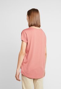 Camiseta de manga corta rosa claro con escote redondo y dobladillo curvo, hecha de una tela suave y lisa. Detalles de costura visibles.