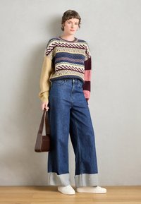 Flerfarget strikket genser med geometriske mønstre, kombinert med vide blå denim bukser med frynset nederkant og hvite sneakers.