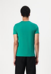 Green cotton t-shirt with short sleeves, a round neck, and a fitted design. Worn with black pants, viewed from the back.