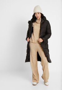 Longue doudoune noire avec des boutons, pull texturé beige, pantalon large beige et baskets blanches. Un bonnet tricoté beige complète le look.
