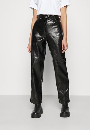 Osoba ubrana w błyszczące czarne, skórzane spodnie z wysokim stanem, czarne masywne botki i prostą białą koszulkę z krótkim rękawem na jednolitym tle.