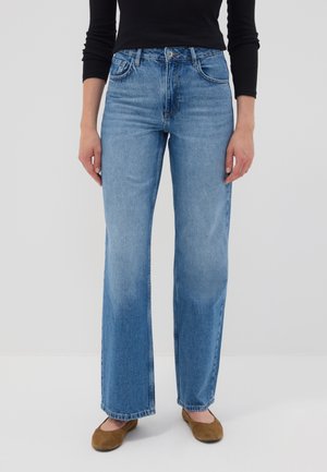 Persona que lleva vaqueros azules de pierna recta, zapatos planos marrones y una camiseta negra de manga larga, de pie contra un fondo liso y claro.