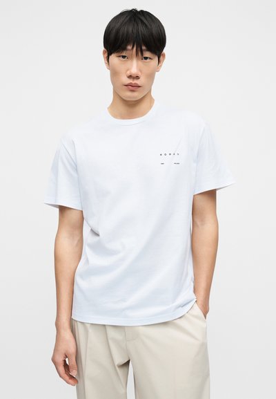 Λευκό βαμβακερό μπλουζάκι με κοντά μανίκια, στρογγυλή λαιμόκοψη και μικρό μαύρο λογότυπο τυπωμένο στο αριστερό μέρος του στήθους. Ομαλή υφή.