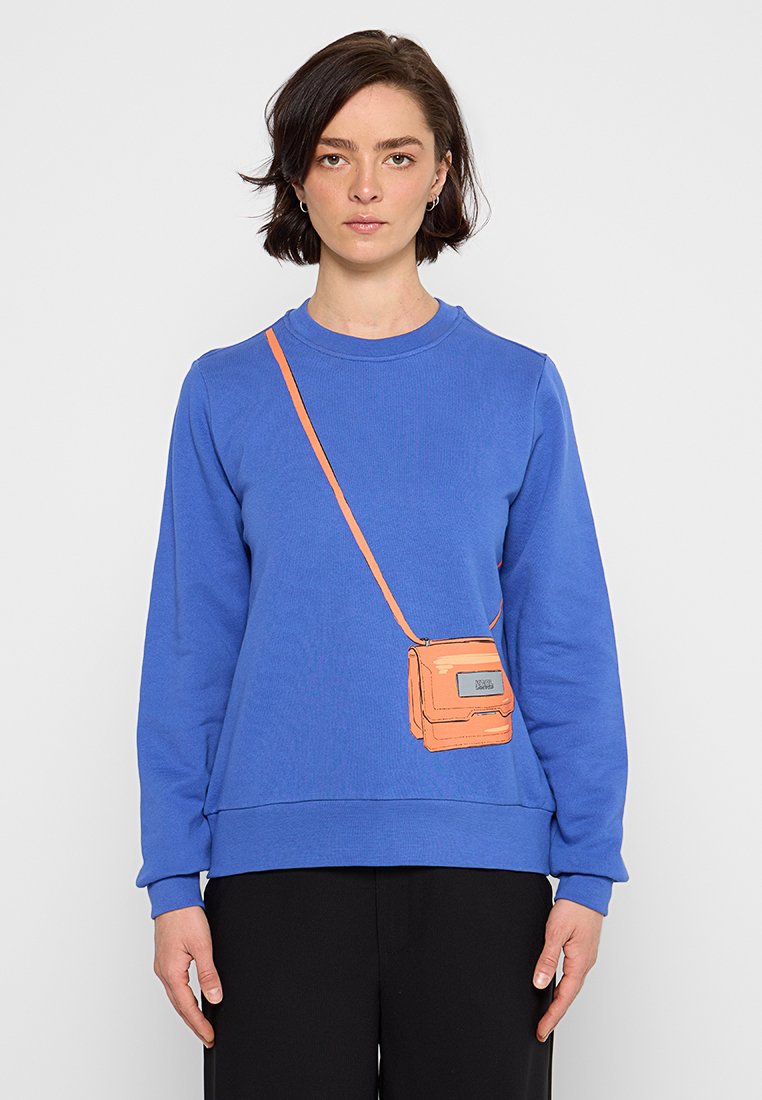Mujer vestida con un sudadera azul y una bolsa cruzada naranja impresa, de pie frente a un fondo claro y liso.